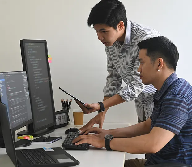 Two engineers working together at a desk with code on the screens