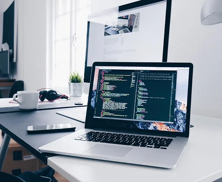 A laptop showing code on a desk, next to a monitor, a phone and a coffee cup